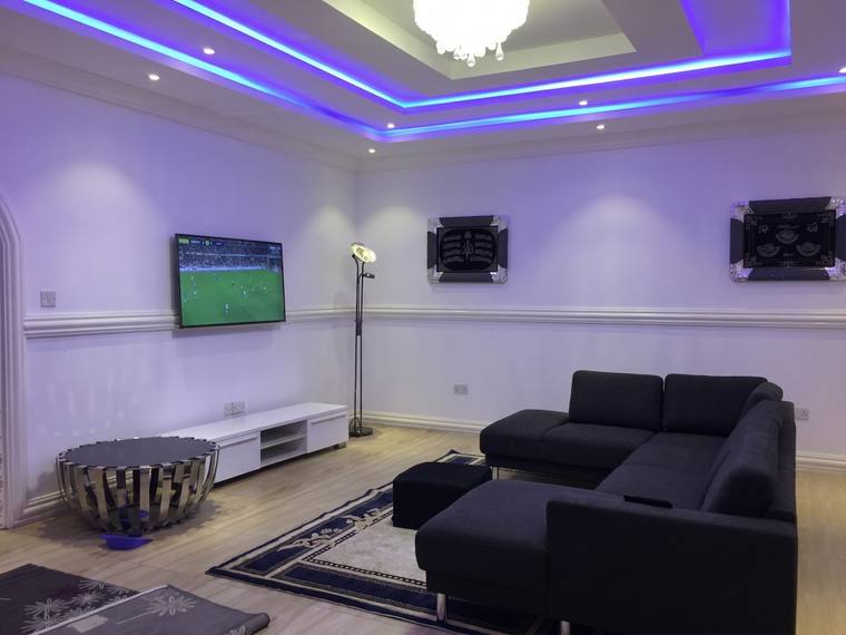 The living room, with a corner sofa, a wall-mounted television and a stepped ceiling carrying cove lighting above plank-effect flooring.
